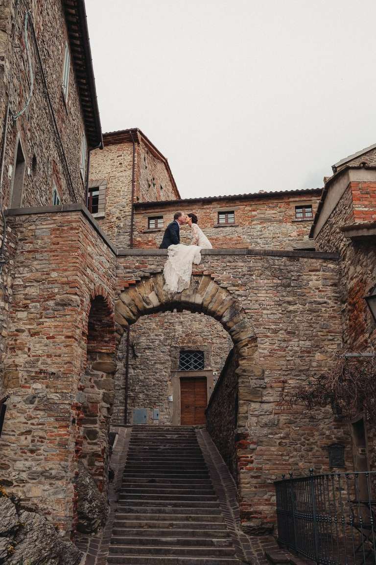 bride and groom sitting atop an old wall in italy